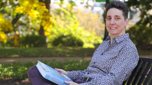Chemistry professor Melanie Nilsson holding a Chemistry textbook while sitting on a bench outdoors.