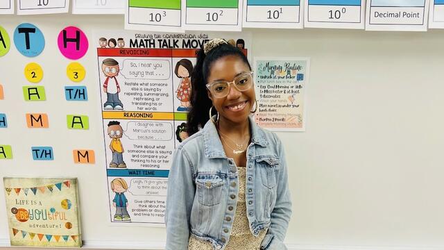Alumna Amara Foster stands in front of a classroom board with colorful art and lessons on it.