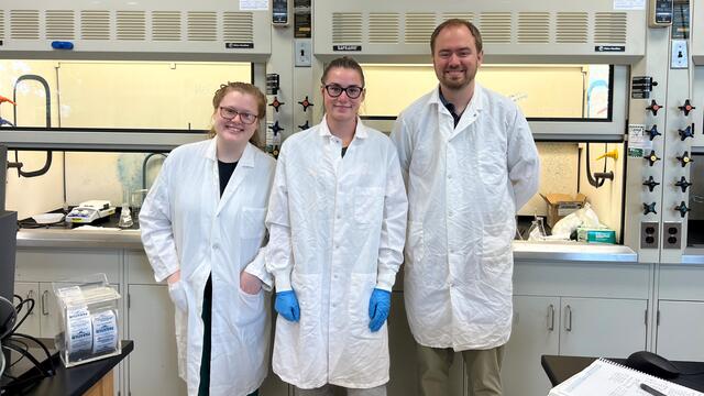 Two students and a professor stand together in the lab, all three wearing white lab coats.