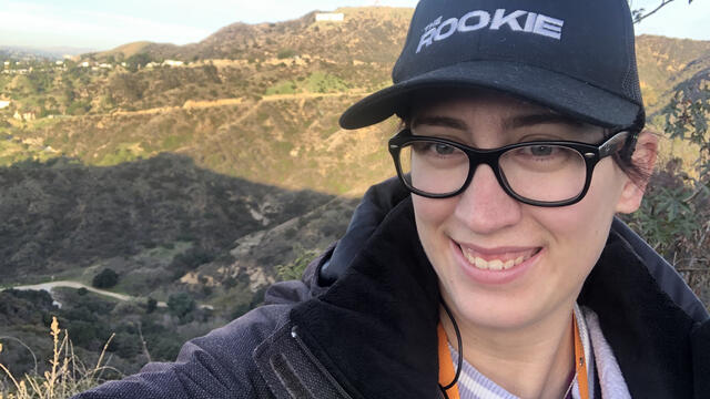 Tricia Meola, a white woman standing in front of the Hollywood sign on a TV set, wearing a hat that says "The Rookie."