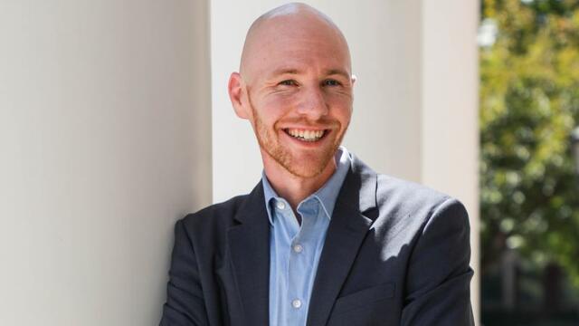 Photo of a white male in a suit jacket and jeans leaning on a white column outside.