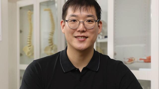 Asian male sits behind a table with anatomy textbooks in front of him and cabinets behind him.