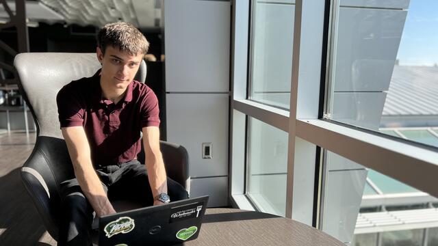 A mal student wearing a black shirt and pants sits by a window with his hands on a laptop.