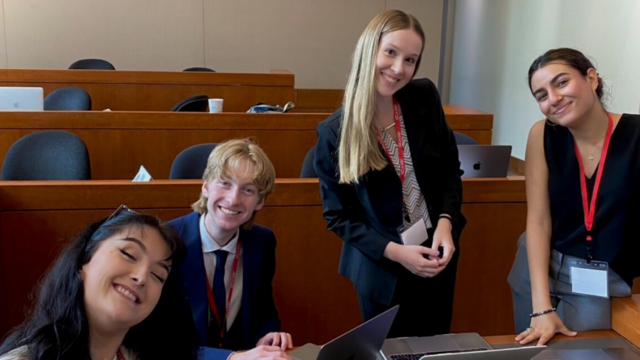 Photo of three female and one male wearing suits and lanyards gathered around a laptop while smiling.