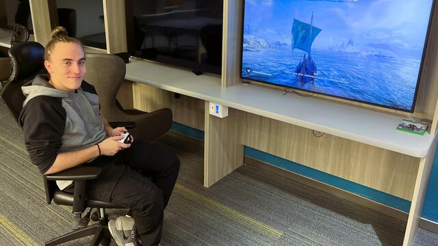 A male student holds a video game controller and smiles at the camera next to a large TV screen with a video game playing on it.