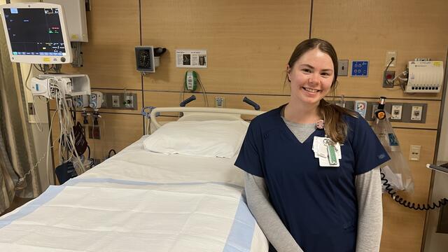 A young woman in nursing scrubs stands next to an empty hospital bed.