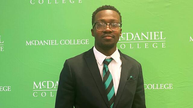 A man wears a suit in front of a green backdrop with the McDaniel College logo on it.