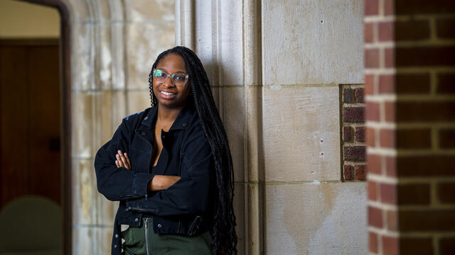 A student stands with arms crossed in a hallways with a stone arch.