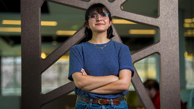 A student stands in front of a metal background with a blue shirt and arms crossed.