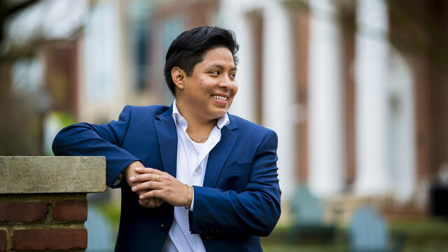A student wearing a suit leans his elbow on a pedestal while smiling off camera.