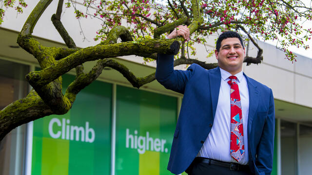 A student in a suit stands in front of a window that reads Climb Higher, and his hand is reaching up to a tree branch.