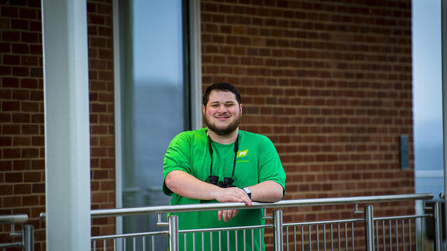 A student leans forward on a railing with a pair of binoculars around his neck.