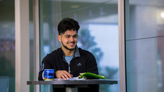 A student sits at a table with a notebook and mug in front of him.