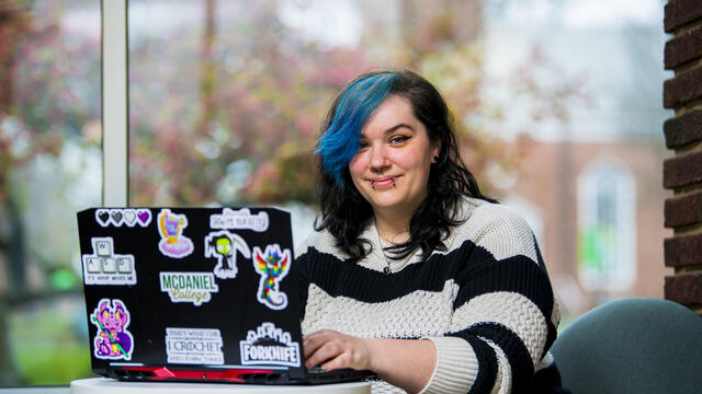 A student in a striped sweater sits in front of a window with a a laptop covered in stickers.