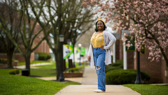 A student in a yellow shirt and grey cardigan stands outside in front of a row of cherry blossom trees.