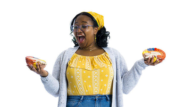 A student in a yellow shirt and jeans holds her hands out to either side while holding two halves of a model brain.