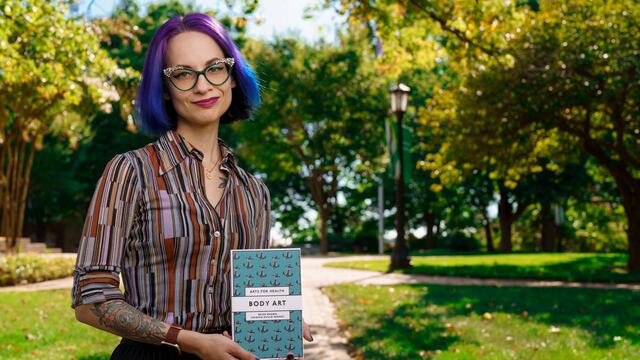 White woman holding a blue book with the title "Body Art" while standing outside.