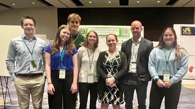 five students and two professors standing by posters at a conference wearing business causal clothes.