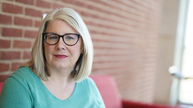 Headshot of Professor Stephanie Madsen with glasses and a blue shirt next to a brick building