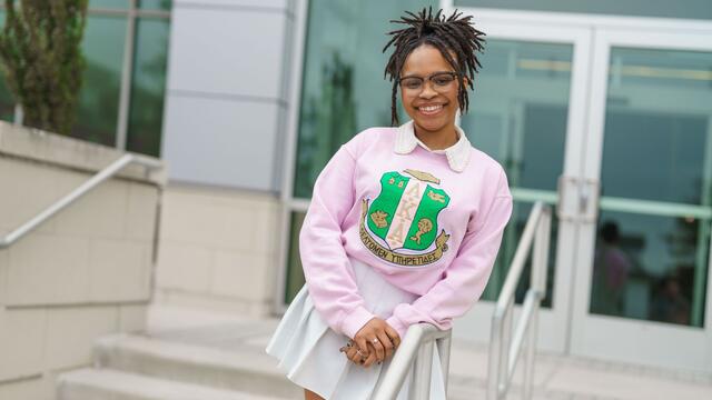 Bria Hill poses outside of the student center in a white pleated skirt and pink sweatshirt.