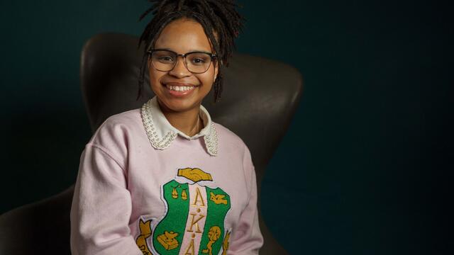 Bria Hill sits in an armchair. She wears a pink sweatshirt with the logo for Alpha Kappa Alpha Sorority.