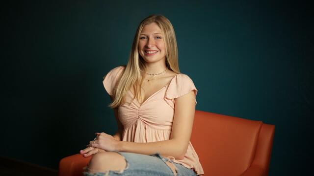 Madge Myers sits on an orange couch. She is wearing a light pink blouse and jeans.