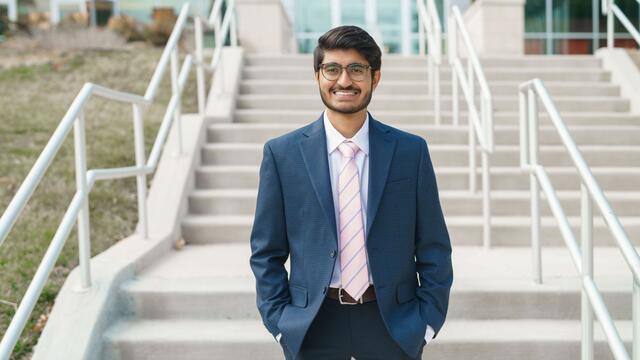 Parth Shah stands outside in front of a set of stairs wearing a navy suit and pink striped tie.