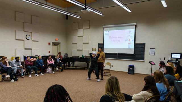 A class of students sit in a semi-circle around two self-defense instructors and a powerpoint screen showing an images labeled "vulnerable areas of the body."