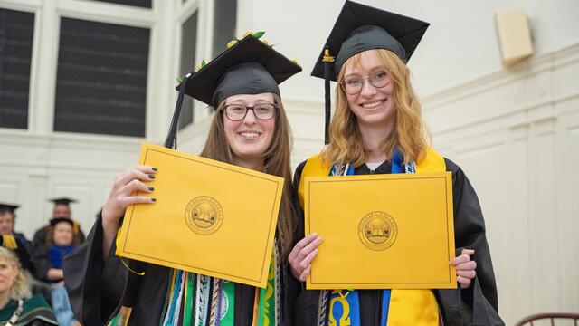 2 students in graduation regalia pose with gold award envelopes