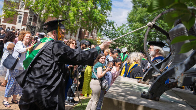 Student in graduation regalia pulls on string to ring large bell with a crowd in the background