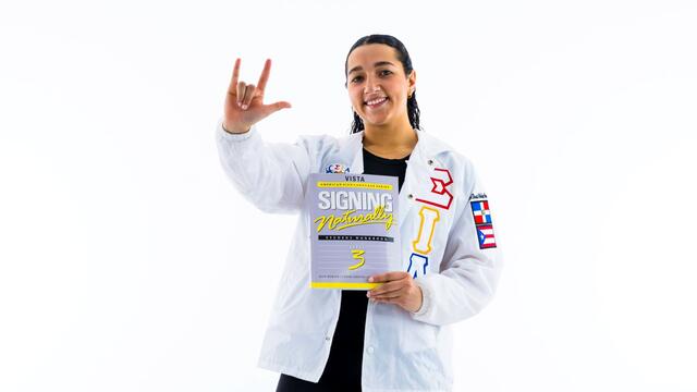 Katherine Taveras against a white backdrop holds up a sign language sign with one hand while holding a textbook on sign language.