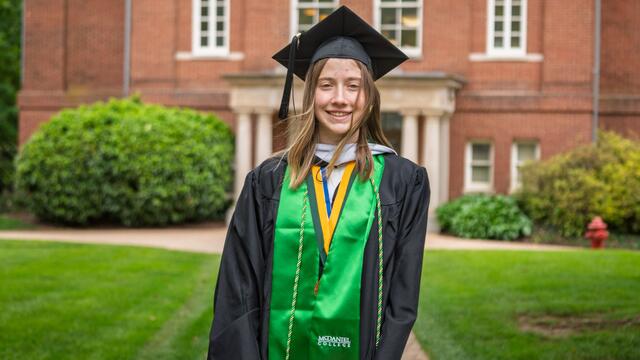 Lorelei McElroy stands in front of a red brick building at McDaniel while wearing her graduation robe and cap with a green stole.