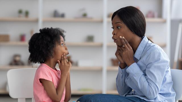 A young woman sits next to a small child at a table. They both hold their hands up to their face while shaping a sound with their mouths.