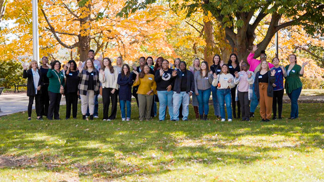 A group of students stand on campus in front of a tree with fall foliage in the background