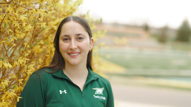 Kate Principio wears a McDaniel track and field jacket and stands in front of a yellow flowering tree.