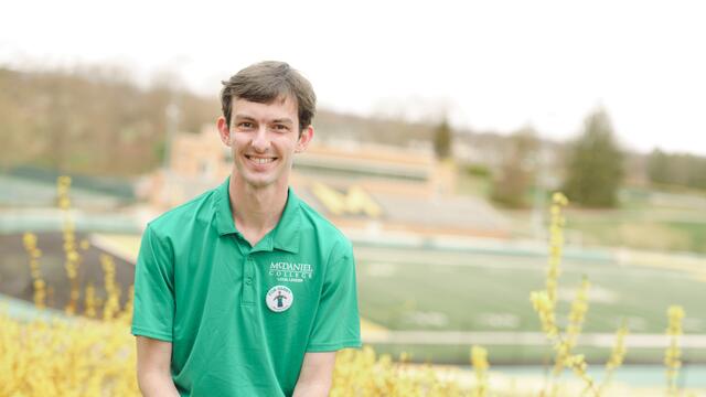 A male student in a McDaniel polo shirt sits in front of the football field.