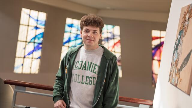 A male student in a McDaniel College shirt stands in front of stained glass windows.