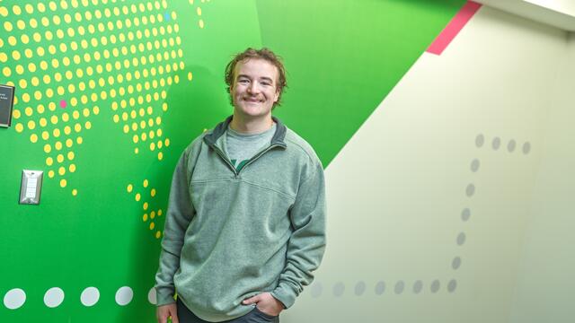 A male student stands in front of a green patterned wall.