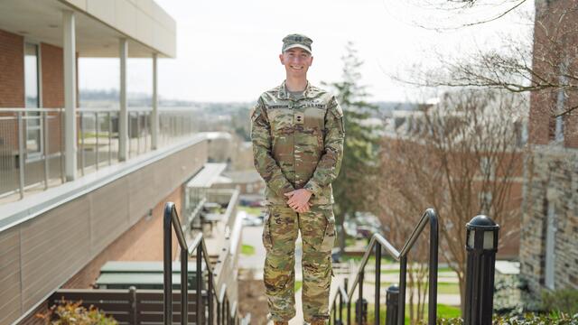 A male student in a military uniform stands on the steps outside of Roj Student Center.