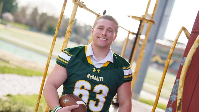 A male student wearing a football jersey smiles and holds a football.
