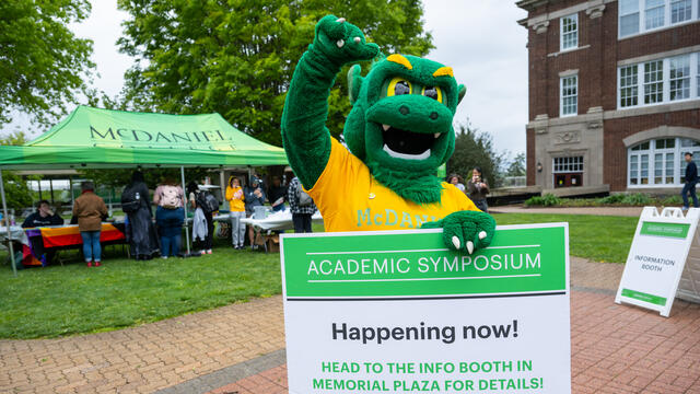 Green Terror mascot holding a sign for the Academic Symposium that reads "Happening Now! Head to the info booth in Memorial Plaza for details!"