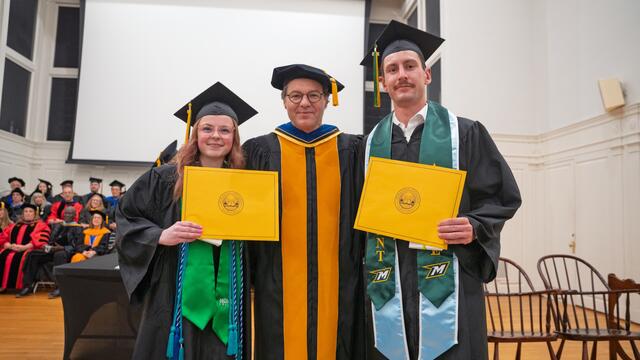 2 students in graduate regalia holding awards stand with the provost in graduation regalia with people seated in the background