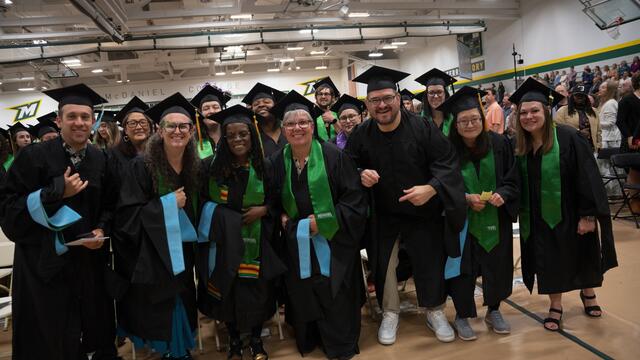 A large group of graduates in caps, gowns, and green McDaniel College stoles pose together smiling in the gymnasium before the graduate commencement ceremony.