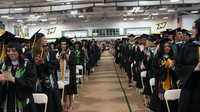 students in graduation regalia stand in rows with an aisle down the middle