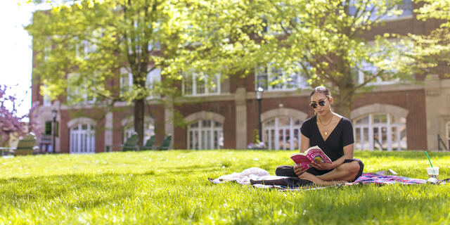 Student reading book sitting on lawn on campus.