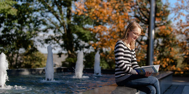 Student reading book sitting on fountain edge on campus.