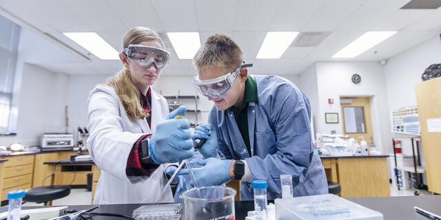 Two students in lab gear create a chemistry solution.