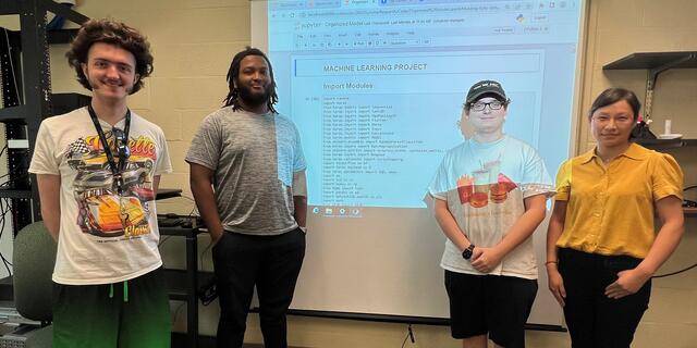 Three students pose with professors Zhang in front of a screen with computer code.