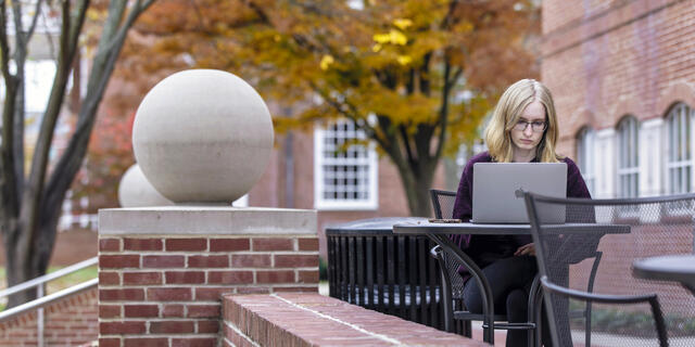 A student sits outside by a brick wall while using her laptop. 
