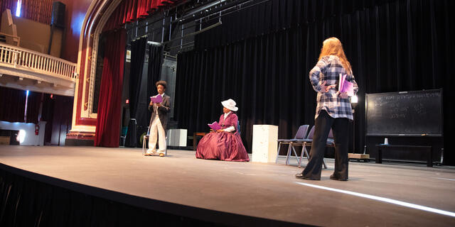 Two students in historical costumes on a stage with a professor directing them.